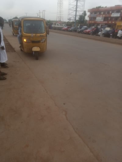 In Tundun Wada South, Nigeria, a yellow auto-rickshaw is parked on a dirt road next to a paved road. The auto-rickshaw is facing left and has its headlights on. A person wearing a white robe and sandals is standing in the foreground to the left of the auto-rickshaw. Several other auto-rickshaws are visible in the background, as well as other vehicles and buildings. The sky is overcast.
