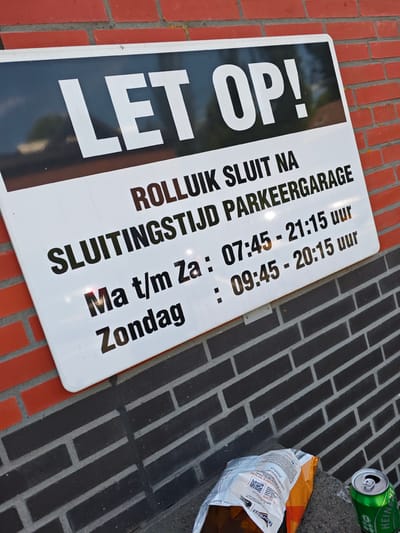 A white sign with black lettering is attached to a red brick wall. The sign reads "LET OP! ROLLUIK SLUIT NA SLUITINGSTIJD PARKEERGARAGE Ma t/m Za : 07:45 - 21:15 uur Zondag : 09:45 - 20:15 uur". Below the sign, on a dark gray brick surface, is an open bag of chips and a green can of Heineken beer. The scene appears to be outside a parking garage in Hoofddorp, Netherlands, based on the Heineken branding.