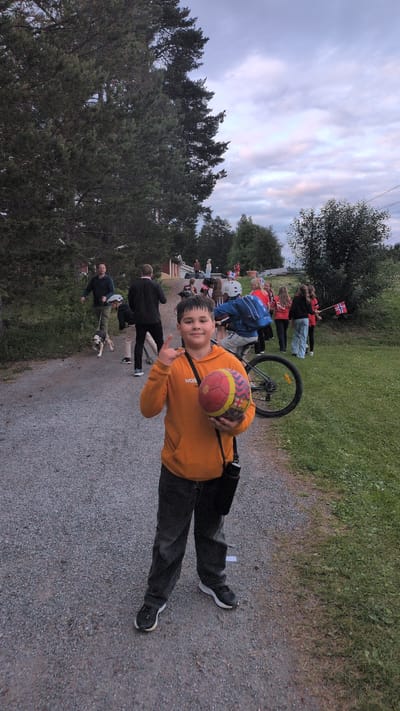 The image depicts an outdoor scene in Östersund, Sweden, likely during the late afternoon or early evening given the soft, diffused light and the color of the sky, which shows streaks of pink and blue in the clouds. The setting appears to be a gravel path bordering a grassy area with trees.
The central subject is a young person, likely a boy, wearing an orange hooded sweatshirt and dark pants. He is holding a colorful ball, possibly for handball, and is making a peace sign with his right hand. He looks towards the camera with a smile, conveying a sense of casual happiness.
In the background, a group of people are gathered, some of whom are holding Norwegian flags, suggesting a patriotic event or celebration. There is a bicycle with its wheel visible, and several individuals are seen walking or standing. The people in the background are dressed casually, with some wearing red and blue clothing. One person is wearing a blue backpack. Further in the distance, a red building is partially visible. The overall atmosphere seems relaxed and festive.
The presence of Norwegian flags strongly suggests a connection to Norway, despite the location being Östersund, Sweden. This could indicate a cross-border event, a celebration related to Norway within Sweden, or simply that the people present are of Norwegian nationality. The presence of a dog being walked also adds to the casual, community feel of the gathering.
The scene is characterized by natural light and an outdoor environment, with trees and grass forming the backdrop. The gravel path suggests a park, a trail, or a recreational area. The overall impression is one of a community gathering, possibly a local event or festival, with a focus on shared enjoyment and celebration.