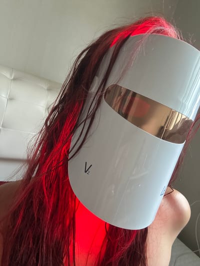 A person with red hair is lying down wearing a white and gold LED light therapy mask. The location cannot be determined.