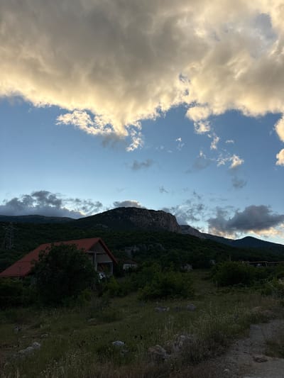 The image depicts a rural landscape with a dramatic sky. In the foreground, a dirt path winds through dry grass and scattered rocks. A house with a prominent orange-tiled roof is situated on the left side of the frame, surrounded by dense green trees. Behind the house, the terrain rises into a series of lush, tree-covered hills. The background is dominated by a large, imposing mountain with a rocky, lighter-colored peak. The sky is a mix of clear blue and voluminous clouds, some of which are illuminated with a warm, golden light, suggesting either sunrise or sunset. A single, tall, metal transmission tower is visible on the far left, partially obscured by trees. The scene appears to be in a relatively undeveloped, mountainous region.