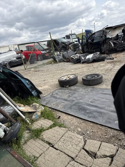 The image depicts an auto salvage yard located in Hrishteni, Bulgaria, under an overcast sky. The ground consists primarily of gravel and dirt, interspersed with patches of grass and broken concrete pavers in the immediate foreground.
In the extreme foreground, a teal-colored car door is visible leaning on the left, near several white metal poles, a shovel, and miscellaneous car parts, including a bare wheel and a brake assembly.
In the mid-ground, two car wheels with tires rest on the ground; one has silver alloy spokes, and the other is a black steel wheel. A large black rubber mat lies on the ground behind these wheels. Beyond this, a green chain-link fence runs across the scene, delineating sections of the yard.
Behind the fence, multiple vehicles are in various states of disassembly and damage. A heavily damaged black minivan or SUV is prominent on the right, with its rear section significantly compromised. A red sedan, partially intact, is visible further left behind the fence. Numerous other vehicle frames and components are scattered throughout this section. In the background, a white semi-truck cab and trailer, along with a blue semi-truck cab, are parked.
One individual, dressed in dark clothing, is standing behind the fence in the mid-ground, positioned between the black minivan and the blue semi-truck. The person appears to be observing or working within the yard.