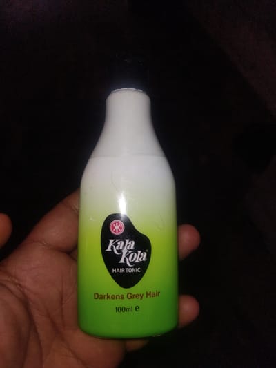 A hand holds a bottle of Kaja Kola hair tonic. The bottle has a white cap and a green body that fades to white at the top. The Kaja Kola logo is visible on the green part of the bottle, along with the text "HAIR TONIC" and "Darkens Grey Hair 100ml e". The background is dark, suggesting low lighting conditions, possibly indoors or at night. The overall scene is simple, focusing on the product. The user's hand is positioned to display the bottle prominently. The location context provided is Haveli Jhangranwali, Pakistan, though there are no specific visual cues in the image to confirm this.