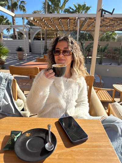 The scene is an outdoor cafe in the Anavissos Municipal Unit, Greece, under a clear, sunny sky. An adult female, with curly brown hair and wearing tortoise-shell sunglasses, a white fluffy zip-up jacket, and distressed light blue jeans, is seated at a wooden table. She holds a black mug containing a light-colored, foamy beverage near her mouth with both hands, looking forward with a slight smile. On the table, to her left, is a black saucer containing light brown residue, a small silver spoon, and a dark green packet. To her right, a dark-colored smartphone rests horizontally. The background features white wooden pergolas with horizontal slats, additional wooden tables and chairs, palm trees, and potted greenery, with distant hills visible under a bright blue sky. Distinct, short shadows indicate direct midday sunlight.