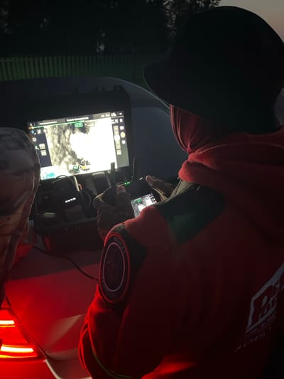 A person, wearing a red hooded jacket and a dark hat, is operating a drone control system. The drone's feed is displayed on a large screen, showing a thermal image of what appears to be a landscape with glowing objects. The control system includes a smaller screen and various buttons and joysticks. The scene is illuminated by the red taillights of a vehicle. The backdrop suggests an outdoor environment with a fence and trees visible in the dimly lit background. The location is identified as дер. Перебор, Russia.