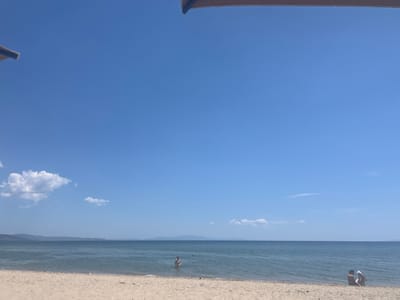 This image depicts a serene beach scene in Nea Moudania, Greece. The foreground features a sandy beach with scattered pebbles. The middle ground is dominated by a calm, blue sea. In the distance, a line of hazy mountains forms the horizon. The sky is a clear, bright blue with a few scattered white clouds.
In the water, two individuals are visible. One person is standing in the shallow water, appearing to be wading. Another, smaller figure is further out in the sea. On the beach, near the shoreline, two people are seated. One is an adult, and the other is a young child wearing a white sun hat. They appear to be interacting.
At the top of the image, a portion of an umbrella or similar canopy is visible, casting a slight shadow. The overall impression is one of a peaceful summer day at the beach.
