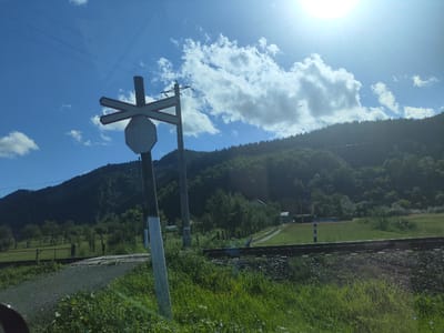 In the city of Leordina, Romania, under a bright sun and partly cloudy sky, a railway crossing sign stands prominently in the foreground. The sign, a post with a white and black striped base, features a white octagonal sign and a red and white "X" shaped crossbar above it. Adjacent to the sign, a single track railway line runs horizontally across the frame, laid on a bed of ballast. Beyond the tracks, a lush green field stretches towards a dense forest that covers a rolling hillside. A dirt road winds through the field, leading to a small building with a white roof. The environment is rural and natural, with power lines crisscrossing the sky.