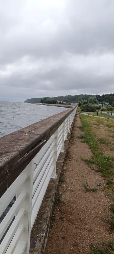 The image shows a wooden and white-railed promenade along a body of water, likely the Volga River in Yuryevets, Russia. The promenade is lined with a sandy path and grass. In the distance, a tree-lined shore with buildings can be seen. The sky is overcast and cloudy, suggesting a gloomy day. There are no people visible in the frame.