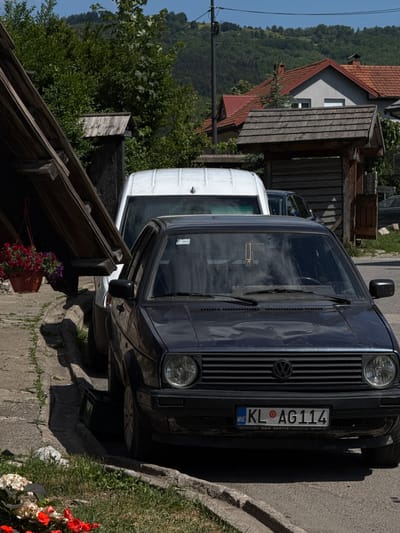 A dark blue Volkswagen Golf Mk2 is parked on the side of a street in Kolašin, Montenegro. The car's license plate reads "KL AG114". A white van is parked behind it. In the foreground, a potted plant with red and purple flowers hangs from a wooden structure. In the background, there are houses with red tile roofs and a lush green hillside under a clear blue sky. The street appears to be paved with asphalt, and a concrete curb runs alongside the parking area. The scene is captured in daylight, with strong shadows indicating a sunny day.