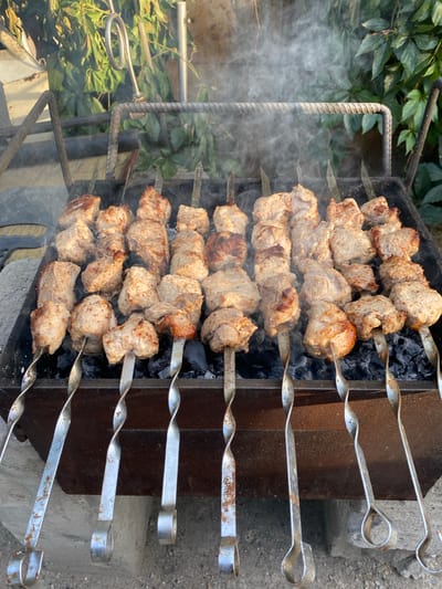 This image depicts a close-up, overhead view of multiple skewers of meat cooking on a grill. The meat appears to be a light brown, suggesting it is pork or chicken, cut into bite-sized cubes and threaded onto metal skewers. The skewers are arranged in rows over hot coals, which are partially visible beneath the meat. Wisps of steam rise from the grill, indicating the cooking process is underway.
The grill itself is a dark metal box, with a rusty, aged appearance. The skewers have twisted handles, designed for easy turning. In the background, lush green foliage is visible, suggesting an outdoor setting. To the right of the grill, a rough concrete surface is present. The lighting suggests it is daytime, likely outdoors. There is no visible text in the image. The overall scene evokes a sense of a casual, outdoor cooking event, possibly a barbecue or a gathering. The location context provided is Әуезов ауылдық округі, Kazakhstan, which places this scene within a rural or semi-rural area of Kazakhstan.