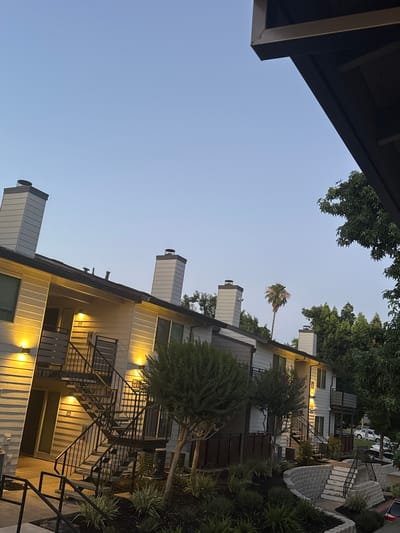 The image depicts the exterior of an apartment building in North Highlands, United States, at dusk. The building is a two-story structure with a light-colored, horizontal siding. It features several external staircases with black metal railings leading to the second-floor apartments. Outdoor lights illuminate the stairs and entrances, casting a warm glow. Two prominent white chimneys with black caps rise from the roof. A few trees, including a tall palm tree, are visible in the background, as well as a portion of a darker, overhang structure in the foreground. The sky is clear and a light blue, indicating the transition from day to night. There are no people visible in the image.