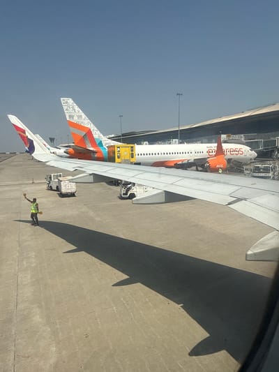 The image shows an airport tarmac from the window of an airplane. The wing of the airplane is visible in the foreground, casting a shadow on the concrete tarmac. Several other airplanes are parked nearby. One airplane has an orange tail with the name "AIR INDIA EXPRESS" printed on its side. A man in a green vest is near the airplane. The location is the Rajiv Gandhi International Airport in Mamidipally, India.
