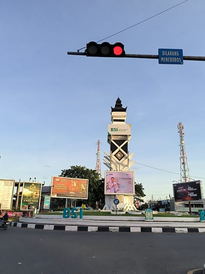 The image depicts a street scene in Lamprehlam Teungoh, Indonesia, likely a town square or a major intersection. Dominating the center of the frame is a tall, white monument with a dark, pointed roof, adorned with geometric patterns and featuring signage for "BSI BANK SYARIAH INDONESIA". Below this monument is a large digital screen displaying an advertisement. To the right of the monument, a red and white telecommunications tower rises into the sky.
In the foreground, a paved road with a distinct black and white striped curb runs across the bottom of the image. A few small pieces of litter are visible on the road surface. To the left, a circular island with grass and landscaping surrounds the monument. Several large billboards are positioned around the island. One billboard to the left shows an advertisement with the text "A MAN TASTE" and a price. Another billboard, located to the left of the monument, features graphics of guitars and text that appears to be an advertisement for "GUDANG GARAM". A large, blue, three-dimensional "BSI" logo is prominently displayed on the circular island in front of the monument.
Above the scene, a traffic light is visible, with the red light illuminated, indicating a stop. A blue rectangular sign with white text that reads "DILARANG MENEROBOS" (meaning "Do not break through" or "No trespassing") is attached to the traffic light pole. The sky is clear and blue with a few faint clouds. A person on a motorcycle is riding on the road in the lower left corner of the image. Another telecommunications tower, taller and with a more industrial appearance, is visible behind the billboards on the left. The overall impression is a modern urban environment with commercial advertising and essential traffic infrastructure.
