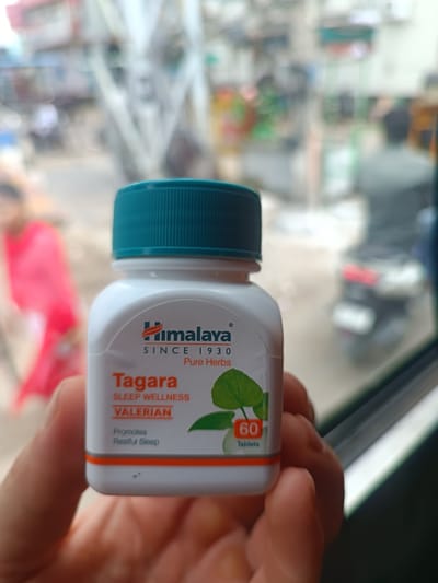 A hand holds a white bottle of Himalaya Tagara Sleep Wellness tablets, with a teal lid. The label on the bottle features the Himalaya logo, "SINCE 1930 PURE HERBS", "Tagara", "SLEEP WELLNESS", "VALERIAN", "Promotes Restful Sleep", and "60 Tablets". The background is blurred, showing a street scene with people and vehicles, suggesting an outdoor or roadside location, possibly in Kurnool, India.