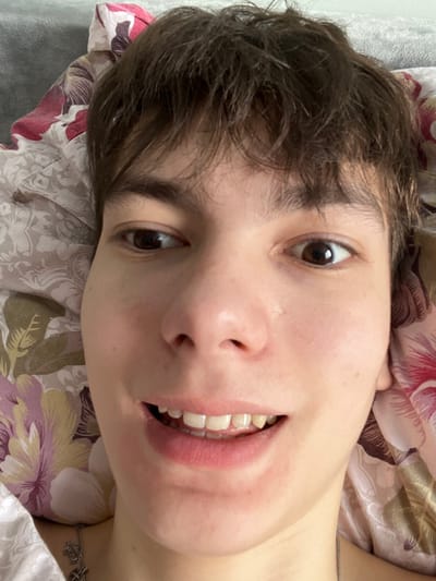 A close-up, high-angle portrait captured indoors in what appears to be a domestic setting within the city of Zainsk, Russia. The image prominently features the face of a single individual, seemingly young, with short, tousled dark brown hair. The person has light skin, dark brown eyes, and is looking towards the camera with a slight smile or smirk revealing both upper and lower front teeth. The individual's left eye (viewer's right) is directed towards the camera, while the right eye (viewer's left) is slightly averted to the side. Visible below the chin, partially obscured by bedding, is a silver-colored chain necklace with small charms, one of which resembles a cross. The person is positioned against bedding featuring a floral pattern in shades of cream, brown, pink, and burgundy. A plain grey surface is visible behind the floral bedding on the upper left.