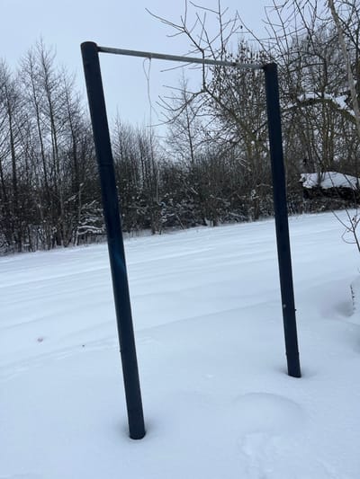 This image captures an outdoor scene in Карабоўскі сельскі Савет, Belarus, under winter conditions. The primary subjects are two dark-colored, cylindrical metal posts, approximately 10-15 cm in diameter, standing vertically from a deep covering of white snow. A lighter grey, cylindrical metal bar, similar in diameter, connects the tops of these two posts horizontally. A single piece of white or light-colored thin material, forming a loose loop, hangs from the left end of the horizontal bar. A faint blue mark or reflection is visible on the upper section of the left vertical post. The ground is uniformly covered in undisturbed white snow, exhibiting subtle undulations. The background features a dense line of bare deciduous trees, indicative of winter, with some light snow visible on their branches and underlying brush. The sky is uniformly light grey and overcast. No human subjects, animals, or active objects are present in the scene.
