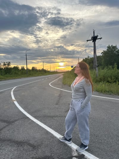 A medium-shot, eye-level, outdoor, full-body photograph shows a Caucasian woman with long, reddish-brown hair standing on an asphalt road during what appears to be sunset. She is facing to the right of the frame, with her head tilted slightly up and her eyes closed, as if enjoying the moment. She is wearing a gray, long-sleeved, wrap-style top with a red necklace, and loose gray sweatpants. Her hands are tucked into the pockets of her sweatpants. She is wearing gray and black athletic shoes.
The road is curved and has white lane markings. The environment is rural, with dense green foliage and trees lining the sides of the road. In the background, a wooden utility pole with electrical wires is visible on the right side of the frame. The sky is filled with dramatic clouds, with the setting sun casting a warm, orange glow on the horizon. The location is identified as Кеткино, Russia.
