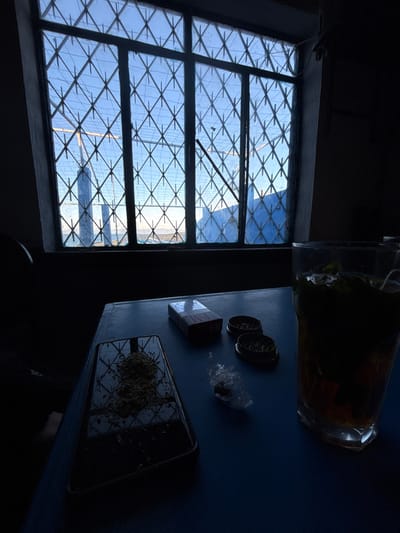 A dimly lit room features a lattice-window, offering a glimpse of a bright, sandy landscape with structures. On a blue table sits a glass of dark liquid, alongside two metallic cylindrical objects, a small wrapped item, and a rectangular object possibly a package. Adjacent to these is a tray containing plant matter. The lighting is uneven, casting significant shadows over the objects and table surface.