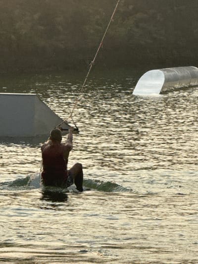 A wakeboarder wearing a maroon shirt is being towed by a cable attached to a watersports structure on a lake. The lake is located in Tambon Nam Phrae, Thailand. The left side of the image shows a dark, forested shoreline. The water reflects the sunlight, creating a shimmering effect.