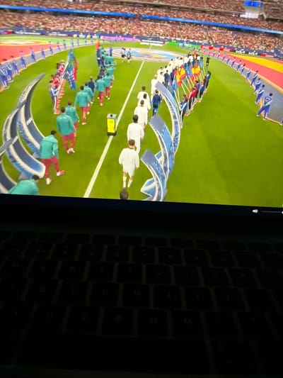 The image displays a soccer match on a large screen, with a laptop keyboard in the foreground. The screen shows players in teal and red uniforms walking in a line, accompanied by individuals holding flags. A stadium with spectators is visible in the background. The laptop keyboard is dark and illuminated by the screen's glow. There is no indication of the city of Nikopol, Bulgaria, in the image.