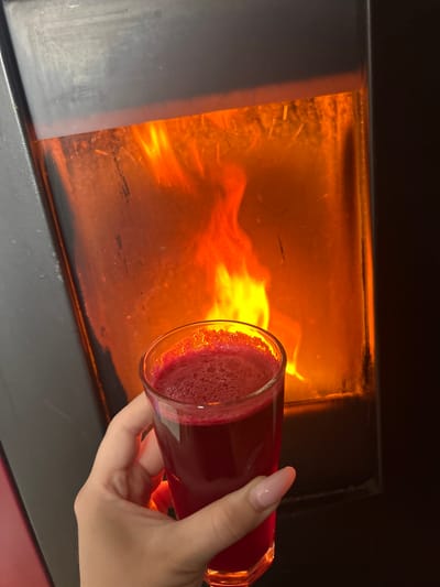 The image depicts an indoor scene within the city of Kumanovo, North Macedonia. The primary subjects are a human hand holding a beverage in front of an active fireplace.

In the background, a dark, rectangular, modern enclosed fireplace or wood-burning stove occupies the majority of the frame. Its central glass panel displays active, bright orange and yellow flames, indicating an ongoing fire. Subtle streaks and residue are visible on the exterior surface of the glass panel, possibly from smoke or heat. The frame of the fireplace is dark, appearing to be black or dark grey metal.

In the foreground, a fair-skinned human right hand grips a clear, faceted glass tumbler. The hand exhibits manicured fingernails with a light pink or nude polish, shaped ovularly. The glass contains a dark red liquid with a frothy layer on its surface, consistent with a freshly prepared juice or similar beverage. The hand is holding the glass steady, presenting the beverage in front of the actively burning fireplace. The ambient lighting is primarily derived from the glow of the flames, with the immediate surroundings of the fireplace appearing dark.