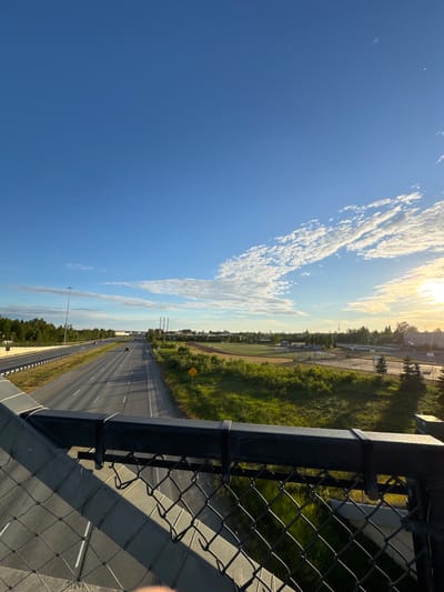 The image shows a highway stretching into the distance under a bright blue sky with scattered white clouds. The sun is setting or rising on the right side of the frame, casting long shadows across the road and surrounding green fields. A chain-link fence and a black railing are in the foreground, suggesting the vantage point is from an overpass. Cars are visible on the highway, moving in both directions. To the right of the highway, there appears to be a sports field or park area. The overall scene depicts a typical suburban or exurban landscape with transportation infrastructure and recreational facilities.