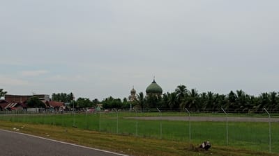 This is a wide-angle, eye-level outdoor photograph of a tropical landscape in Keupok Nibong, Indonesia. The sky is overcast with pale gray clouds. In the mid-ground, a chain-link fence runs horizontally across the frame, separating a grassy field from a paved area. Beyond the fence, a cluster of buildings with red-tiled roofs are visible on the left, some appearing to be homes with corrugated metal extensions. Further to the right, two domed structures, likely mosques, stand out. The larger dome is green, while the smaller one is a lighter color with ornate details. Palm trees are abundant throughout the scene, particularly in the background, forming a dense green canopy. A paved road occupies the lower left portion of the image. Scattered debris, including a piece of what appears to be a damaged barrel, is visible in the grass near the fence.