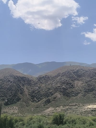 This image captures a wide, sweeping landscape of rolling mountains under a bright, clear blue sky with scattered white clouds. The foreground features a rugged, rocky mountainside covered in sparse, dry vegetation, with some areas appearing eroded. Further back, more distant mountain ranges are visible, appearing softer and more muted due to atmospheric haze. A thin, dark vertical line on the far left might indicate a utility pole or a signpost.
On the crest of one of the mid-ground mountains, a tall, slender structure, likely a telecommunications tower, is visible. The overall scene suggests a natural, remote, and somewhat arid environment. The time of day appears to be daytime, with bright sunlight casting shadows and illuminating the landscape. The weather is fair and clear. The location context provided, Kok Moynok Eki, Kyrgyzstan, aligns with the mountainous and potentially arid terrain depicted. There is no visible text in the image. The mood conveyed is one of vastness and natural grandeur.