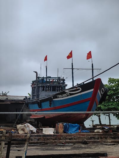 A large, blue and red-hulled fishing vessel, identified by the registration "TTH94899TS" on its black bow, is dry-docked on elevated wooden supports in Thuận An Ward, Vietnam. Three red Vietnamese flags with yellow stars fly from its masts, one forward and two aft. The vessel features a grey/blue multi-deck superstructure with windows and an exhaust pipe. Black cylindrical fenders are visible on the bow. The ground beneath and around the vessel is an uneven surface of dirt and debris, including scattered wood, concrete blocks, white sheeting, blue tarpaulins covering objects, and empty barrels. A metal ladder leans against the port side of the vessel. A single human figure is indistinctly visible near the ladder on the vessel's port side. In the background, a low building with a corrugated metal roof is partially visible to the left, while green foliage from trees lines both sides. A white, cylindrical beacon is faintly visible in the far background to the right. The sky is uniformly overcast and grey.