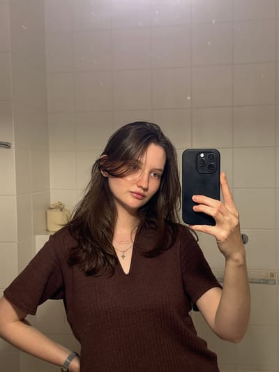 A young woman with long, dark brown hair is taking a selfie in a bathroom. She is wearing a dark brown, short-sleeved shirt and a silver necklace with a cross pendant. Her left hand is on her hip, and her right hand is holding a black smartphone, positioned to capture her reflection in the mirror. The bathroom features white tiled walls and a tiled floor. A roll of toilet paper is visible on a shelf to the left of the woman. The lighting appears to be artificial. The specific location of Журавлево (кемеровский), Russia, cannot be determined from the visual information provided in the image.