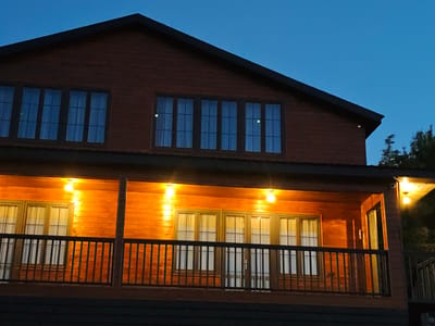 A wooden house with a dark roof and dark brown siding is illuminated by outdoor lights. The house has multiple windows with a grid pattern. On the lower level, a balcony with a black railing overlooks the front of the house. Two lights are visible on the balcony, casting a warm glow on the wooden exterior. The sky is a deep blue, indicating it is evening or night. Trees are visible on the right side of the image. The image appears to be taken from a slightly low angle, looking up at the house. The location is identified as Ukeshi, Georgia.