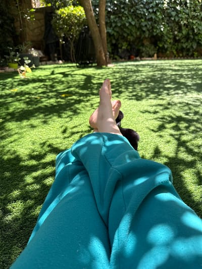A close-up, outdoor shot shows a person sitting on artificial grass, possibly in a yard or garden. The individual is wearing blue-green sweatpants. Part of a foot is visible on the left side. The green artificial grass is dappled with shadows, probably from trees or bushes. A glimpse of foliage and branches can be seen in the background on the left. The location is not discernible as Ariel, Palestinian Territories from the visual elements in the image.