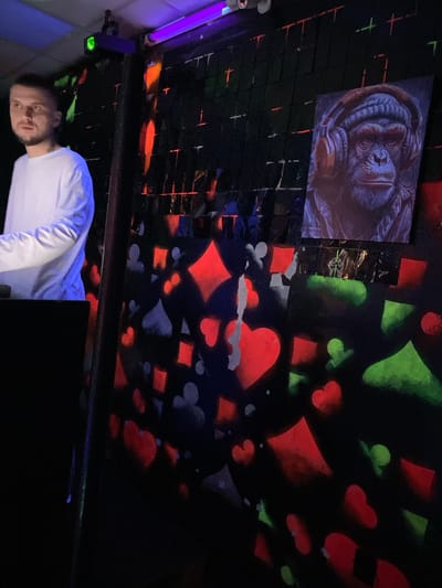 In Kizner, Russia, within a dimly lit indoor venue adorned with neon or blacklights, a man stands to the left, facing forward and slightly to the right. He has short, dark hair and a beard, and is wearing a long-sleeved white shirt. He appears to be engaged with something out of frame to his left, possibly a DJ booth or a control panel, as his posture suggests interaction with equipment.

The background wall is decorated with a grid of dark squares, some of which emit faint neon or blacklight illumination in red, green, and orange, creating a vibrant, abstract pattern. Several playing card suit symbols, specifically diamonds, hearts, and spades, are depicted in neon red and green spray-paint or stencil art across the wall, adding to the energetic atmosphere.

Mounted on the wall to the right is a framed artwork, a portrait of a chimpanzee wearing headphones and a beanie. The artwork is illuminated, contrasting with the darker elements of the background. The overall environment suggests a club, entertainment venue, or a themed party space.