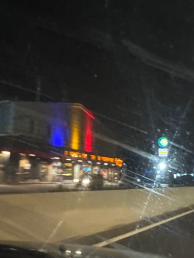 The image presents a nighttime scene in Jilava, Romania, viewed through a vehicle's streaked or smudged windshield. The primary subject is a two-story commercial building on the left. Its upper facade is distinctly illuminated by three prominent vertical light panels in blue, yellow, and red, aligning with the colors of the Romanian national flag. The ground floor features multiple brightly lit windows, revealing an interior with visible shelving or merchandise, indicative of a retail establishment or supermarket, partially obscured by a dark red awning extending along its front.
A blurred vehicle with active headlights is visible on the street directly in front of this building. To the right, a tall pole-mounted commercial sign stands, displaying a circular green logo with a white internal design, positioned above a rectangular sign with orange lettering, consistent with the branding of a Mega Image supermarket. The foreground consists of a paved road with at least one discernible white lane marking. The overall environment is dark, with artificial lighting sources providing illumination. No individuals are distinctly visible within the frame.