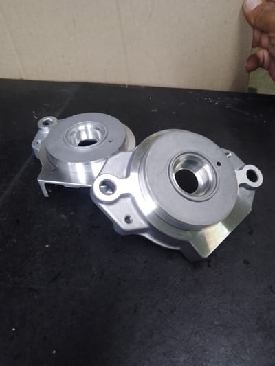 The image shows two metallic objects on a dark, textured surface, likely a table or workbench. The objects are identical, appearing to be some type of mechanical component or housing, possibly for machinery or automotive parts. They are made of a polished, silver-toned metal, with intricate machining evident. Each object features a circular opening in the center, with raised lips and internal detailing. There are also mounting holes and projecting flanges.

The background is a mottled, light brown surface, possibly a wall or partition, with a hint of a cardboard-like texture. In the upper right corner, a person's hand is partially visible, with fingers extended towards the background surface, suggesting someone is holding or interacting with it.

The lighting is somewhat diffused, creating highlights and shadows on the metallic objects, which emphasize their three-dimensional form and machined surfaces. There are some small white specks and smudges on the dark surface, indicating a workshop or industrial setting. The overall impression is of manufactured parts ready for assembly or inspection. No text is visible in the image. The scene appears to be indoors, in what could be an industrial or manufacturing environment in Ciantra, Indonesia.