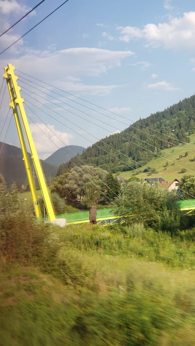 The image depicts a suspension bridge with a distinctive bright yellow pylon and green deck, spanning across a body of water. The bridge is situated in a picturesque natural setting with lush green hills and dense forests in the background. A statue of a bear stands on the bridge, adding a unique element to the scene. The sky is a clear blue with a few scattered clouds. The overall impression is one of a serene and visually striking landscape. This bridge is located in Švošov, Slovakia.
