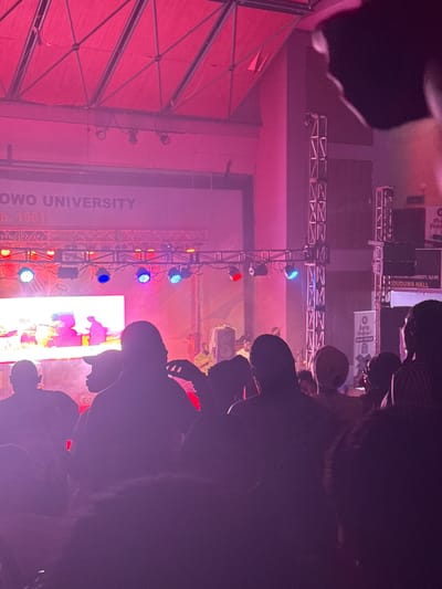 The image captures an indoor event, likely a concert, performance, or university gathering, viewed from the audience's perspective. The scene is dominated by vibrant stage lighting and a large display screen.

In the foreground and to the right, numerous individuals are visible in silhouette, appearing as an audience or attendees, observing the activity on stage. Their bodies are mostly dark, with subtle outlines and occasional highlights from the stage lights.

The main focus is the stage area, illuminated by an array of red, blue, and white stage lights mounted on a metal truss structure. A prominent large white screen on the stage displays bright, colorful, and possibly abstract graphics or video content.

Several banners are suspended above and behind the stage. Visible text on these banners provides significant location cues: "OWO UNIVERSITY," "b. 1961," "UNIVERSITY, ILE-IFE," and "ODUDUWA HALL." These details strongly indicate the event is taking place at Obafemi Awolowo University in Ile-Ife, Nigeria, within Oduduwa Hall, which was established in 1961. Other partially visible text includes "Proplev" and "Prab."

The overall atmosphere suggests an evening or night event, given the reliance on artificial stage lighting. The combination of colorful lights, a large screen, and an engaged audience indicates a dynamic and lively gathering within a university setting.