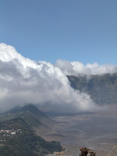 The image shows a panoramic view of a volcanic landscape with rolling hills, a vast plain, and a dramatic cloudy sky. In the foreground, a lush green hillside slopes down towards the right, dotted with trees and a cluster of buildings that appear to be part of a town or resort. A winding road is visible on the plain in the middle ground. The background features a barren, sandy plain stretching towards distant, hazy mountains. Clouds are a prominent element, partially obscuring the mountain peaks and creating a sense of depth and atmosphere. The sky above is a clear blue. The location is identified as Cemorolawang, Indonesia.