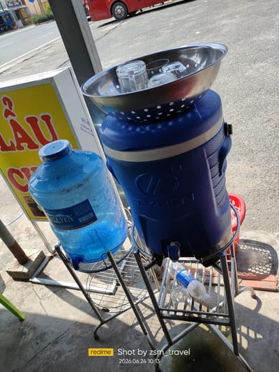 In Gò Dầu, Vietnam, an outdoor scene depicts a beverage stand situated on a concrete surface. The focus is on two large blue water dispensers and a metal stand supporting them. The dispenser on the left is a large, clear plastic bottle filled with blue-tinted water, condensation visible on its surface. The dispenser on the right is a larger, opaque blue container with a dispenser spout at its base. A stainless steel bowl containing several clear glasses rests on top of this dispenser. Below the dispensers, a metal shelf holds a partially filled plastic water bottle and another glass. A yellow sign with red lettering is partially visible behind the left dispenser, and a red bus is seen in the background on a paved road. The lighting suggests a sunny day, casting shadows on the ground.