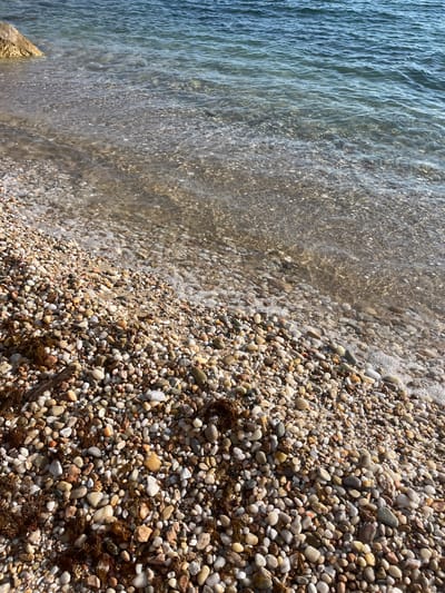 The image shows a pebble beach with clear, shallow water lapping at the shore. The pebbles are of various sizes and colors, predominantly white, grey, and brown, with some interspersed reddish-brown seaweed. A large rock is visible in the upper left corner of the image, partially submerged in the water. The water is a deep blue and appears calm with gentle ripples. The scene is likely a coastline in Potos, Greece, characterized by its rocky beaches and clear Aegean Sea. There are no people or identifiable man-made objects in the frame.