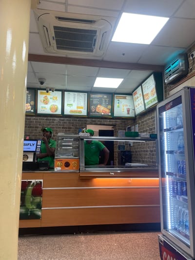 The image shows the interior of a Subway restaurant in Gollapalle Khurd, India. Two men, likely employees, are visible behind the counter, one in a green uniform with a cap and the other partially visible, also in a green uniform. They appear to be working, with one taking an order on a phone and the other possibly preparing food.

The setting is typical of a fast-food establishment, with menu boards displayed on the wall showing prices and food items. Visible text on the menu boards includes "HOT & CHEESY," "WRAP," "SALADS & WRAPS," "FRESH SALADS," "FOOTLONG," and "Cookies." There are also advertisements for "SIFLY" and "CAMPA." In the background, a refrigerated display case contains bottled beverages. An air conditioning unit is mounted on the wall, and a security camera is visible.

The lighting suggests it could be daytime or evening, as the interior is well-lit with overhead fluorescent lights. The overall atmosphere appears to be functional and busy, consistent with a food service environment.