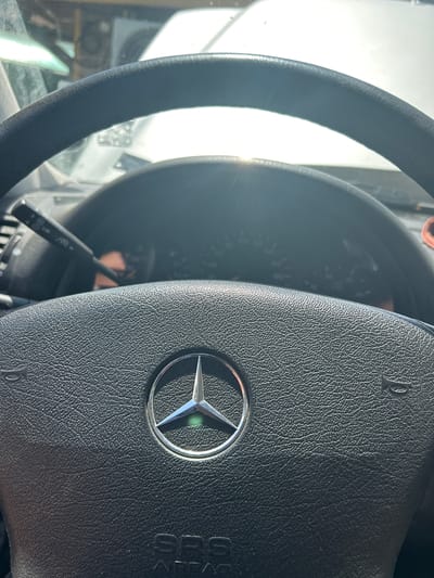 This is a close-up, high-angle shot of the interior of a Mercedes-Benz vehicle, focusing on the steering wheel and dashboard. The steering wheel is black, with a textured leather grip and a silver Mercedes-Benz emblem in the center. To the right of the emblem, the word "SRS" is visible, indicating an airbag. The dashboard is mostly out of focus, but a speedometer with markings up to 200 can be partially seen, along with a windshield wiper stalk. The bright sunlight streaming in from the left suggests it is daytime. The scene is likely inside a car parked or being driven, given the interior view. There are no people visible in the frame. The overall impression is one of a functional car interior bathed in natural light. The location context provided is Dulovo, Bulgaria.
