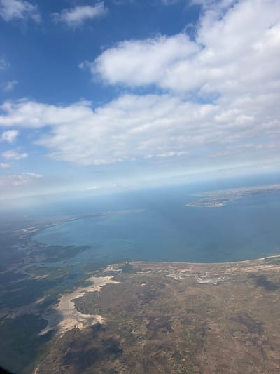 This media file is an aerial photograph taken from an airplane, looking down at a coastline. The image is oriented vertically, giving it a tilted perspective. The scene depicts a transition from a bright blue sky filled with white, wispy clouds on the left to a calm, dark blue ocean on the right.

The ocean meets a landmass characterized by a sandy, light-colored coastline that curves inland. Beyond the coastline, the land appears to be a mix of scrubland, sparse vegetation in shades of brown and green, and darker patches that could be shadows or denser foliage. There are visible waterways or inlets from the sea that snake into the land. In the distance, along the coastline, there are hints of what might be settlements or infrastructure, appearing as scattered lighter-colored patches and linear features.

The time of day appears to be daytime, possibly late morning or early afternoon, given the bright sunlight and the shadows cast on the land. The weather seems clear, with the sun shining through the scattered clouds. The location appears to be a coastal region with a distinct transition from sea to land, and the terrain suggests a relatively dry climate with pockets of vegetation.

There is no visible text in the image. The overall impression is of a vast, natural landscape viewed from a high altitude, highlighting the geographical features of a coastal area.