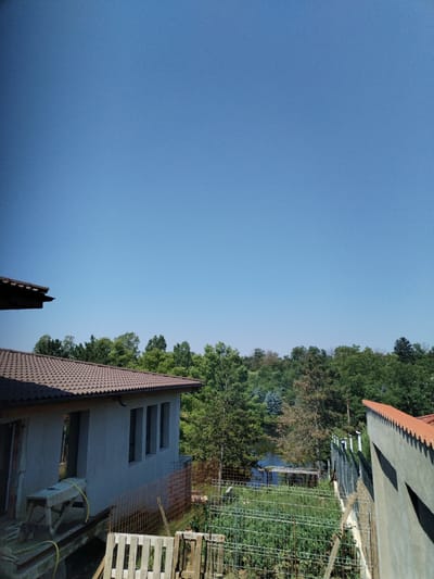 The image depicts a clear, blue sky dominating the upper half. Below, a treeline of varying shades of green forms a horizon. In the foreground, a light gray, unfinished building with a brown tile roof is visible on the left. The building has three rectangular windows and a partially constructed awning. In front of the building, there is a makeshift wooden table with a yellow hose draped over it. To the right of this building, a fenced-in garden area contains rows of green plants, likely vegetables. Further to the right, another building with a similar brown tile roof is partially visible, casting a shadow on its exterior wall. A fence separates the garden from the foreground, and beyond the trees, a small body of water is visible. The scene appears to be a rural or suburban setting, potentially a residential property under construction or renovation, in Moara Domnească, Romania. There are no people visible in the image.