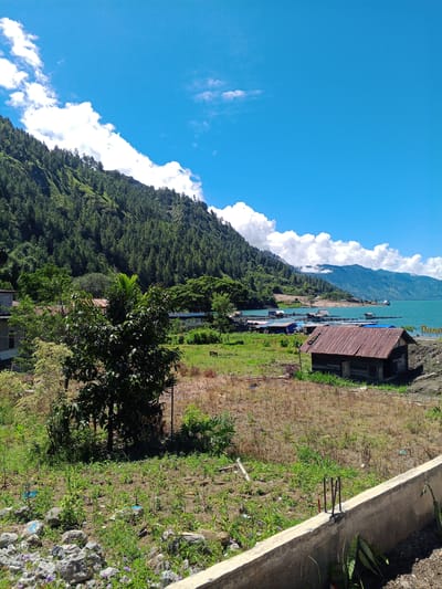 This is a wide-angle, high-angle, natural-light, outdoor photograph of a scenic landscape in Mendale, Indonesia. The scene captures a clear, bright blue sky with scattered white clouds. A lush green, tree-covered mountainside dominates the left side of the image, sloping down towards a body of turquoise water. In the middle ground, there are several small, rustic buildings and structures situated near the water's edge, some appearing to be over the water on stilts, suggesting a village or a collection of fish farms. A prominent, weathered, wooden shack with a rusted metal roof is positioned in the foreground on the right. The ground in the foreground is uneven and covered with dry grass, scattered rocks, and some patchy green vegetation. A concrete wall runs along the very bottom edge of the frame, with rebar protruding from it. The overall impression is one of a rural, waterside community nestled against a dramatic natural backdrop.