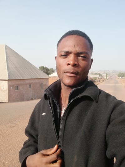 A man stands outdoors wearing a black zip-up jacket with a collar. He is unzipping the jacket with his left hand. A building with a pyramid-shaped roof is visible in the background to his left. A road extends into the distance behind him, lined with buildings. The sky is clear and pale. He is wearing a small necklace with a cross on it.