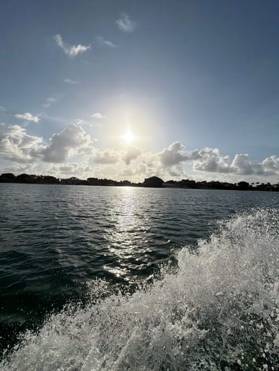The image shows a scene from the water, likely a bay or inlet, near North Miami, Florida. The view is oriented vertically, with the sky and clouds on the left, and the water on the right. The sun is visible through the clouds, creating a bright glare. A landmass with trees and some buildings is situated between the water and the sky. The water is dark and rippled, with sunlight reflecting off the surface. On the right edge of the water, there is a splash or wave. It's unclear if this is caused by a boat moving through the water or natural wave action. No people are visible.
