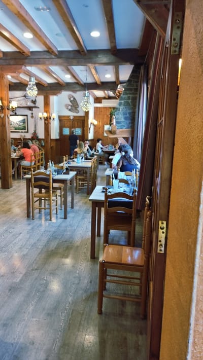 The image depicts the interior of a restaurant in Setcases, Spain, characterized by a rustic, wooden aesthetic. The ceiling features exposed wooden beams and recessed lighting. Several tables are set with chairs, some occupied by patrons, while others are empty. The flooring appears to be a light-colored wood laminate. On the left side of the image, patrons are seated at tables, some engaged in conversation, and others with their backs to the camera. In the background, a fireplace is visible, constructed of stone. The right side of the image is dominated by a wooden doorframe and a textured wall. The overall ambiance suggests a casual dining establishment.