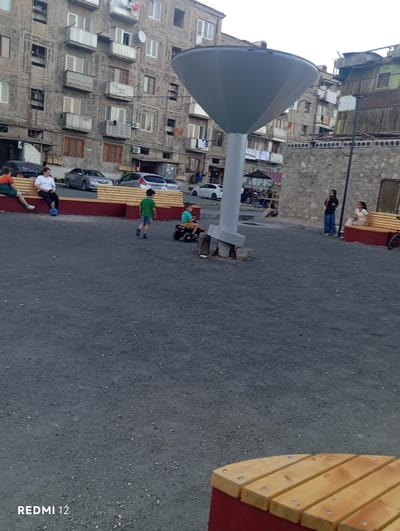 This media file depicts an outdoor scene in Vedi, Armenia, likely in the late afternoon or early evening, given the soft lighting and lack of harsh shadows. The setting appears to be a public space adjacent to residential buildings, possibly a courtyard or park area.

The primary subjects are several people engaged in various activities. Two individuals are sitting on a long, wooden bench with a red base. One person, wearing a white t-shirt and dark pants, is reclining on the bench, while another, wearing a green t-shirt and dark shorts, is standing and looking towards the bench. Further along the bench, another person in a black jacket and blue jeans stands, facing away from the camera. In the background, a child in a green shirt is pushing a small toy or cart.

The scene also features several residential buildings with a distinctive architectural style, characterized by multiple stories, numerous windows, and balconies. Some buildings appear to be older, with weathered facades and some visible wear and tear. The ground is paved with asphalt, typical of urban environments. A large, grey, parabolic-shaped object, possibly part of a playground or public art installation, dominates the left side of the frame. A red and yellow decorative wooden structure, likely part of a bench or play area, is visible on the right. Several cars are parked along the side of the paved area, suggesting it might also serve as a parking space.

There is no discernible text visible in the image, except for "REDMI 12" in the bottom right corner, which is likely a watermark from the device used to capture the photo. The overall atmosphere appears relaxed and casual, with people enjoying the outdoor space. The weather seems to be overcast or the light is diffused, as there are no strong highlights or deep shadows.