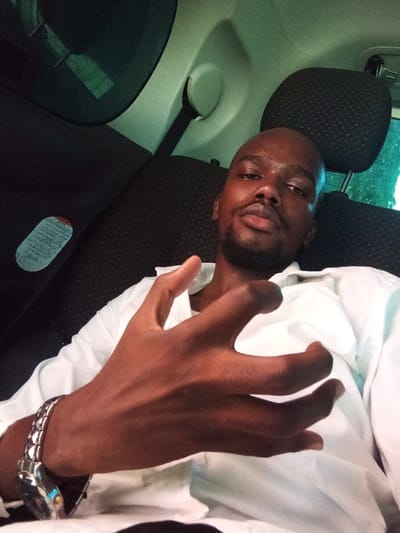 A dark-skinned male, bald with a short beard, is seated in the interior of a vehicle, in what is identified as Ganjoni/Shimanzi ward, Kenya. He wears a white collared shirt and a silver-toned wristwatch on his left wrist. His left hand is raised in front of him with fingers spread, while his gaze is directed towards the camera. The vehicle's interior features dark fabric seats and lighter-colored trim. A dark-tinted window is visible on the left side, showing faint green foliage outside. A sticker with Japanese text is affixed to the vehicle's interior panel below the window. Another dark headrest is visible behind the subject to his right.